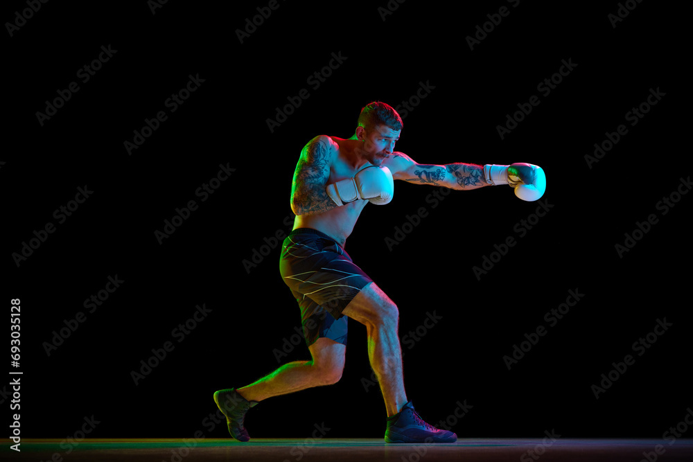 Young muscular man, shirtless boxing athlete in motion, training, practicing hooks isolated over black background in neon light. Concept of professional sport, combat sport, martial arts, strength