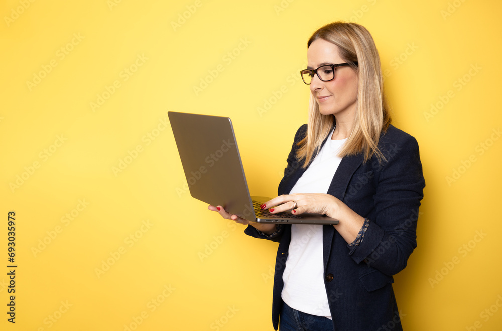 Smiling cheerful funny beautiful attractive young woman 40s wearing basic casual jacket standing working on laptop pc computer isolated on yellow colour background studio portrait Smiling cheerful funny beautiful attractive young woman 40s wearing basic casual jacket standing working on laptop pc computer isolated on yellow colour background studio portrait