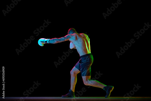 Young muscular man, shirtless boxing athlete in motion, training, practicing hooks isolated over black background in neon light. Concept of professional sport, combat sport, martial arts, strength