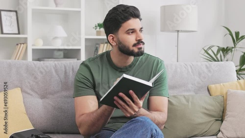 Handsome bearded guy using personal copybook for noting thoughts while sitting on couch and looking pensively aside. Indian man in casual attire enjoying free time alone at home with new insights. photo