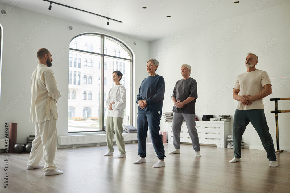 Wide angle shot of senior people standing in gym and doing qigong exercise for energy concentration while male master guiding them - obrazy, fototapety, plakaty 