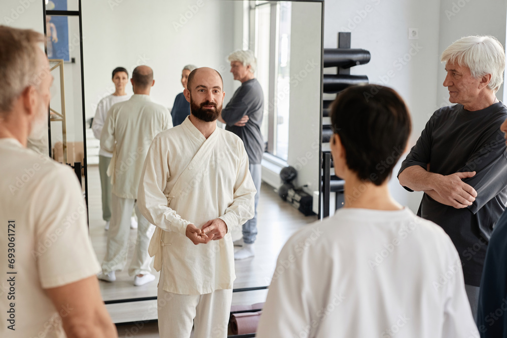 Obraz premium Wide shot of bearded trainer in white apparel explaining exercise programme to senior qigong class participants in gym