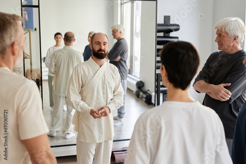 Wide shot of bearded trainer in white apparel explaining exercise programme to senior qigong class participants in gym photo