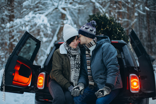 A couple poses in the trunk of their car with a Christmas tree on the roof in a winter forest.