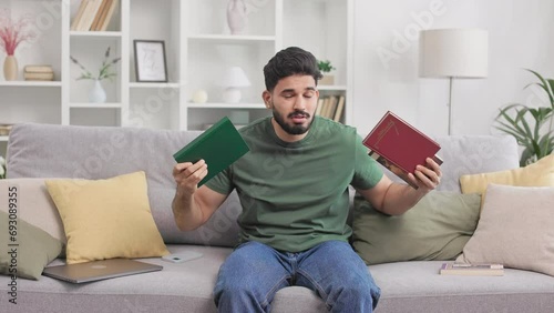 Indian male student feeling stressed of hometask while sitting on couch and surrounded with books. MIllennial guy in casual attire touching head with hands during difficulties before exams. photo