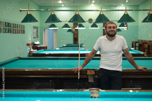 A happy handsome man stands in the billiard room, leaning on the table and chatting with friends. The man is having a blast having fun with his friends and playing billiards.