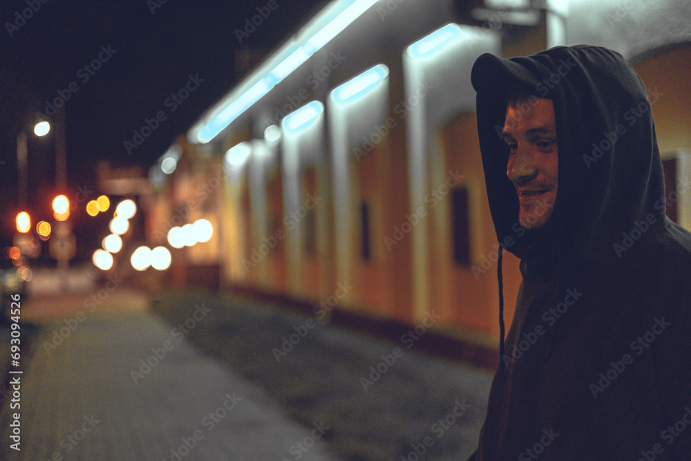 A handsome man in the neon light of night city signs. A man with a hood over his head walks through the city at night. Neon lights from the signs illuminate a hooded man. A handsome man in the neon light of night city signs. A man with a hood over his head walks through the city at night. Neon lights from the signs illuminate a hooded man.