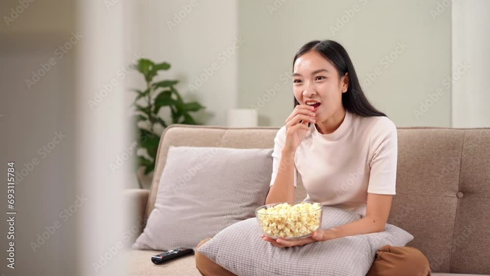 Young asian woman watching funny movie TV on sofa something excited in the film, looking and laughing eating popcorn glass bowl snack at living room, Mass media and emotion concept Young asian woman watching funny movie TV on sofa something excited in the film, looking and laughing eating popcorn glass bowl snack at living room, Mass media and emotion concept