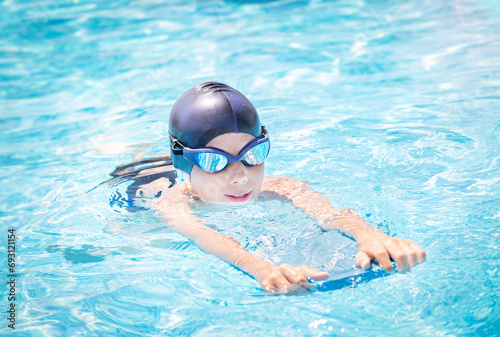 Active child (boy) in cap, sport goggles doing water sport with swim board in the swimming pool. Healthy lifestyle.