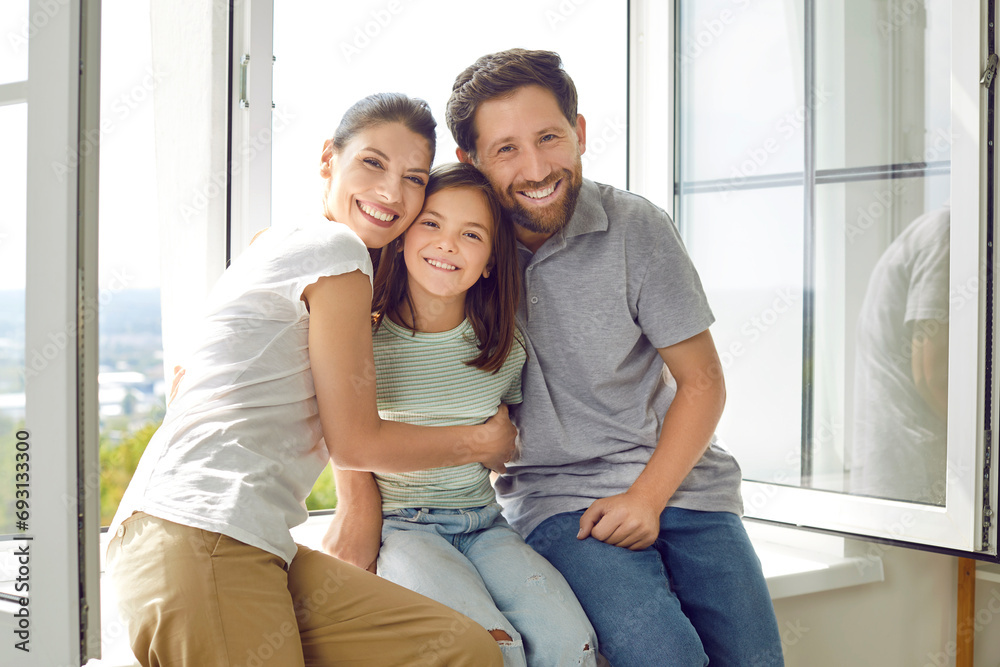 Portrait of smiling young happy family of three with daughter sitting on the windowsill at home and looking cheerful at camera hugging their child girl. Love, care, family time and leisure concept. Portrait of smiling young happy family of three with daughter sitting on the windowsill at home and looking cheerful at camera hugging their child girl. Love, care, family time and leisure concept.