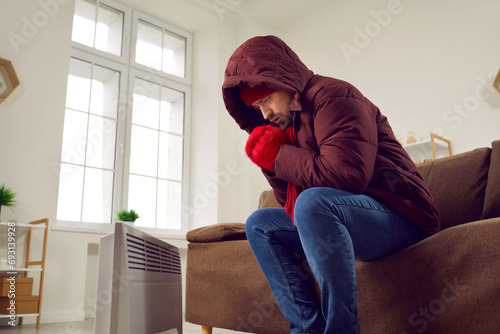 Unhappy young man sitting on couch feeling cold at home. Shivering man wearing winter jacket, knitted hat and gloves warming himself at electric heater feeling cold at home at cold winter season © Studio Romantic