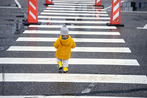 Stylish kid toddler girl in yellow rubber boots and coat on a striped pedestrian crossing on the road alone photo
