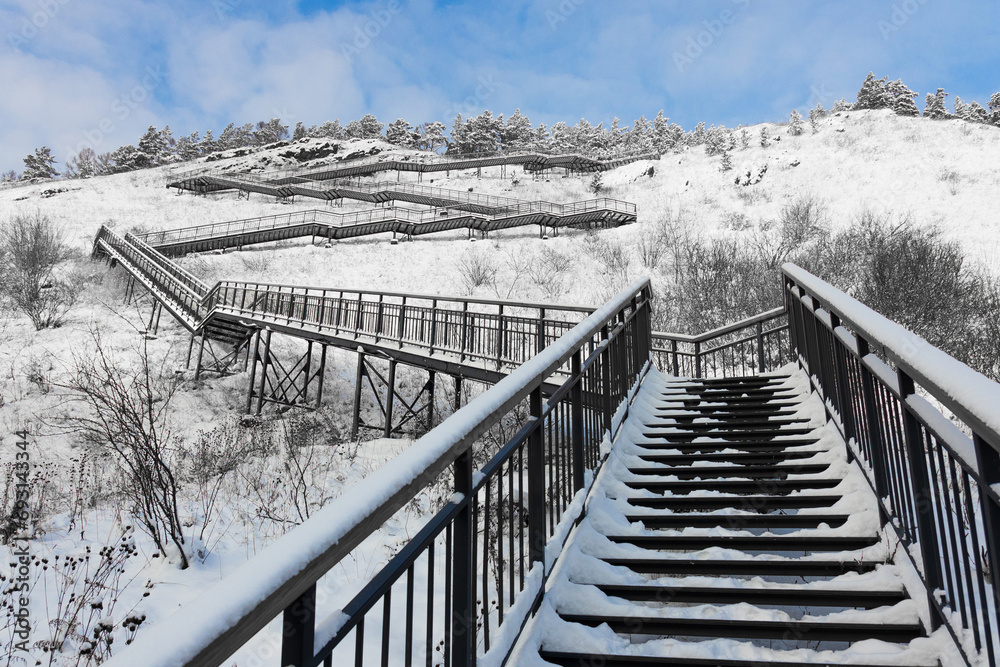 Snow-covered staircase with metal steps curves along the hillside at Krasnoyarsk, Russia. Winter landscape with steps, snow-covered pine trees on top and blue sky. Snow-covered staircase with metal steps curves along the hillside at Krasnoyarsk, Russia. Winter landscape with steps, snow-covered pine trees on top and blue sky.