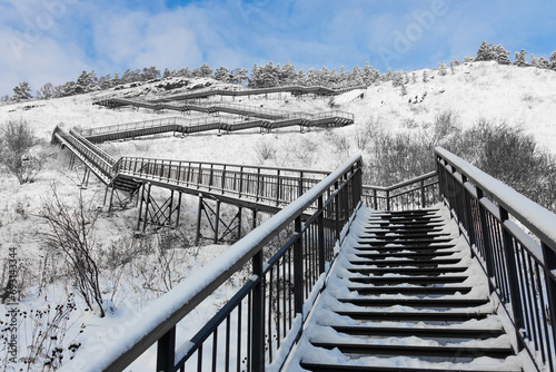 Snow-covered staircase with metal steps curves along the hillside at Krasnoyarsk, Russia. Winter landscape with steps, snow-covered pine trees on top and blue sky.