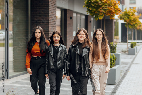 Group of smiling and happy teenage friends wearing casual clothes spending time together, posing and talking with each other near college building on autumn day.