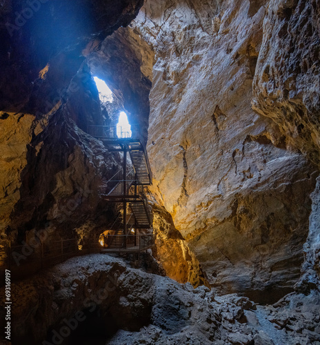 Ancient cave interior with a metal staircase leading towards a sunlit opening above. photo