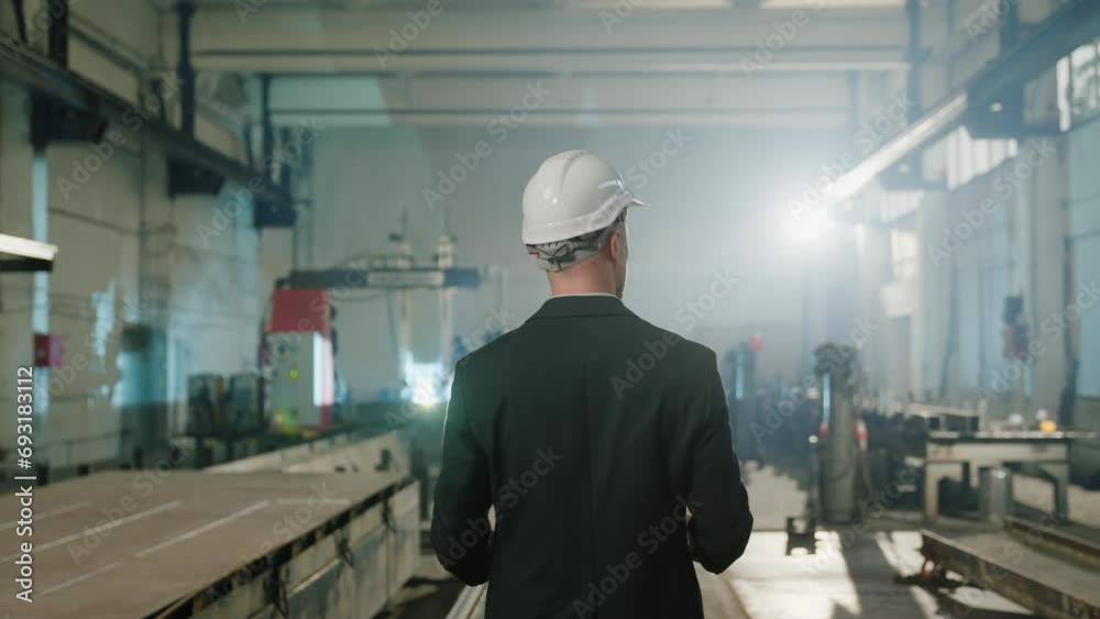 In front of the camera taking video from the back of main manager with safety helmet he holding the plan of work and standing in the middle of manufacturing production zone In front of the camera taking video from the back of main manager with safety helmet he holding the plan of work and standing in the middle of manufacturing production zone
