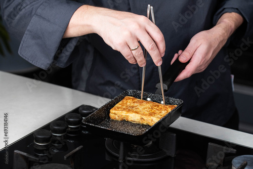 Chef man hands cooking Tamagoyaki or Tamago traditional Japanese Rolled Omelette recipe with chopsticks, made by rolling several layers of cooked scrambled egg in pan into a rectangular omelet. photo