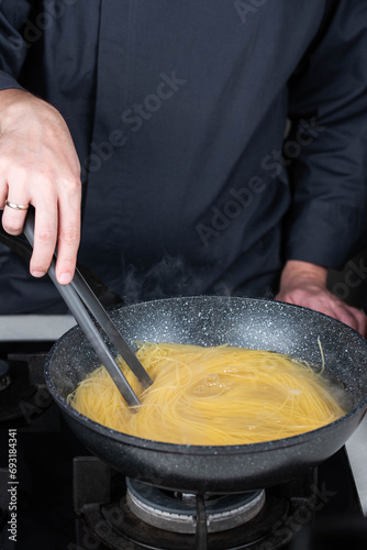Professional chef cooks making Italian Capellini spaghetti pasta at modern kitchen gas stove in wok pan pot in boiling water. Steam from hot food. 