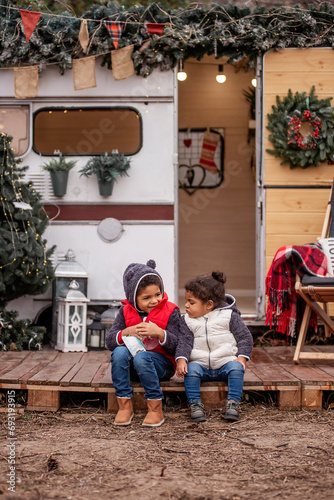 African American boy and girl are sitting at the camper on wooden pallets, playing with a Christmas snow globe. Brother and sister shake the ball, snow flies in it. Christmas holidays outside the city © farmuty