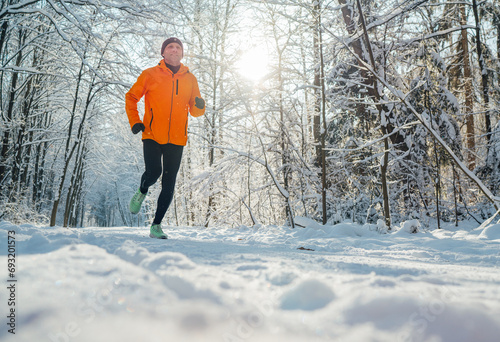 Middle-aged smiling trail runner man dressed bright orange windproof jacket endurance fast running picturesque snowy forest during sunny frosty day. Sporty active people and winter training image.