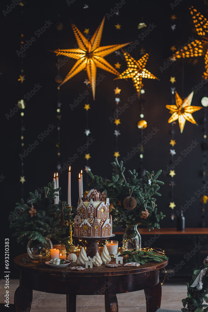 Fototapeta premium Christmas and New Year beautiful elegant family dinner table setting decorated with candles, fir tree branches, handmade craft toys. Gorgeous big homemade cake with gingerbread houses as centrepiece.