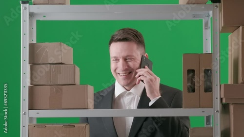 In the frame on a green background. Depicts an adult man in a suit. Demonstrates the manager, the boss in a warehouse. He checks boxes talking on the phone and smiles joyfully. Medium frame photo