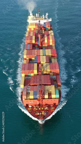 Container Ship underway calm waters with a dolphin surfing the bow wave. Vessel with white trails all around on nice deep green shade of ocean. Complete deck extends from prow to aft. Aerial vertical