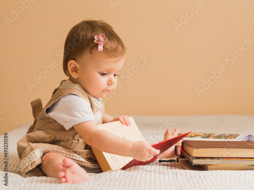 Portrait of 1 year cute baby girl in beige dress sitting on a sofa with stack of books and holds a red book in her hands	
