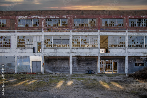 Facade of a derelict former factory building with broken windows, missing roof, streaks of sun in foreground, dark sky, nobody