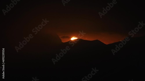Il sole scivola dietro lo skyline nero delle montagne al tramonto, sullo sfondo di un cielo rosso e arancione fra banchi di nebbia photo