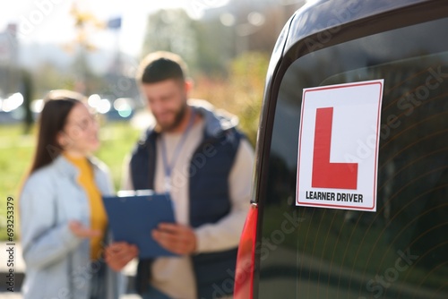 Learner driver and instructor with clipboard near car outdoors, selective focus on L-plate. Driving school photo