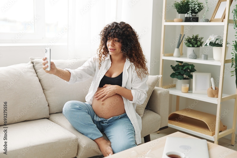 Pregnant woman blogger sits on the couch at home and takes pictures of herself on the phone, selfie and video call, consultation with the doctor online, pregnancy management Pregnant woman blogger sits on the couch at home and takes pictures of herself on the phone, selfie and video call, consultation with the doctor online, pregnancy management