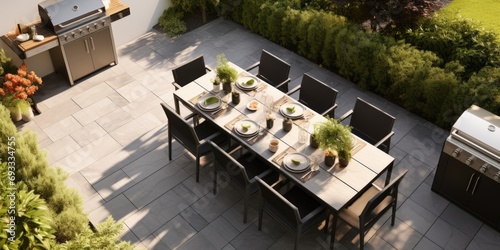 Stylish outdoor kitchen with gas barbecue and dining table set for guests, formal place settings and flowers on paved patio, seen from high angle. photo