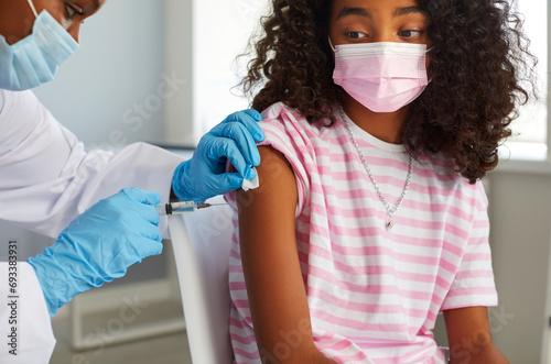 Doctor african american woman is giving vaccine injection to teen girl in shoulder in clinic. Healthcare, vaccination from flu, covid-19, immunization concept. They wearing protective medical masks. photo