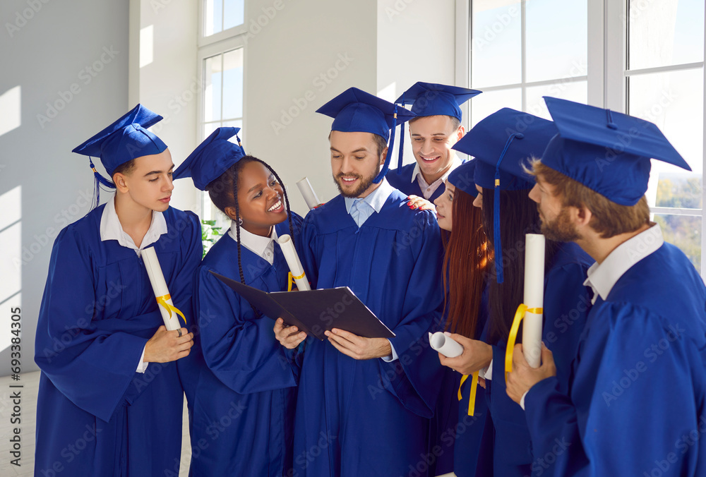 Happy smiling diverse multiethnic graduate students in blue graduation robes celebrate graduation and looking to diploma of their classmate standing indoors. Education and graduation concept. Happy smiling diverse multiethnic graduate students in blue graduation robes celebrate graduation and looking to diploma of their classmate standing indoors. Education and graduation concept.