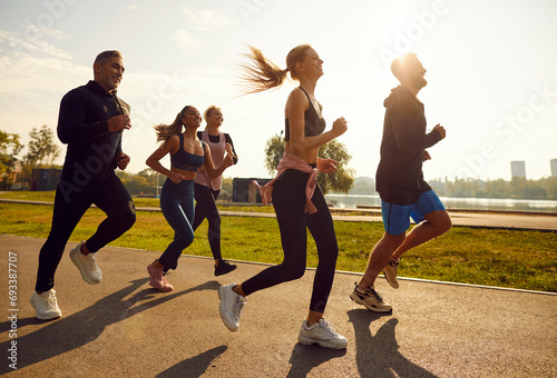 Group of sporty smiling people in sportswear jogging together in the park at sunset. Friends running outdoor having sport training in nature. Team of runners on workout. Sport and fitness concept. #693387707