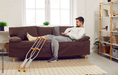 Handicapped young man lying at home on sofa in rehabilitation with his crutches and working on a laptop. Brunet guy in rehab after injury with injured leg typing on computer or chatting with friends.