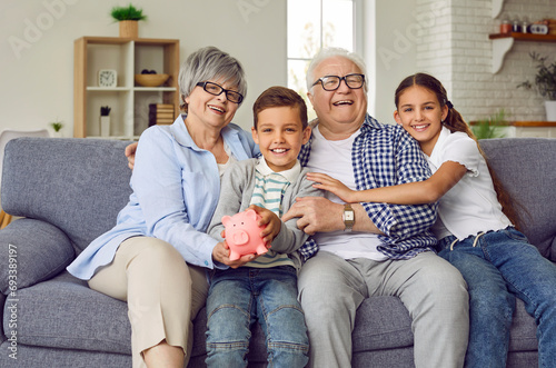 Cuadro en lienzo Portrait of a happy grandparents sitting on sofa with their grandchildren boy and girl in the living room at home looking at camera and holding piggy bank in hands
