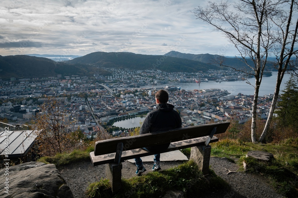 Traveler sitting on a bench, with his back turned, enjoying a panoramic view from a viewpoint of the city of Bergen, Norway. Traveler sitting on a bench, with his back turned, enjoying a panoramic view from a viewpoint of the city of Bergen, Norway.