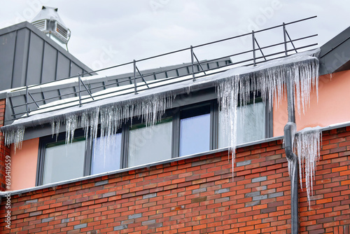 Icicles hanging on roof of modern residential building. Sharp icicles hang from edge of the roof. Icicles above the entrance, risk to life. Icicle on drain pipe of the building.