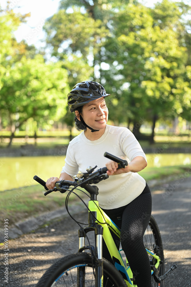 Fototapeta premium Portrait of happy retired woman in helmet riding bicycle in park on sunny summer day.