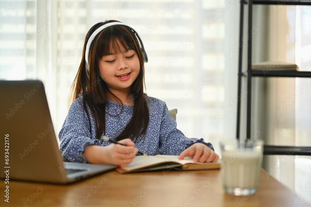 Smiling Asian schoolgirl in headphone studying online on laptop, doing homework at dinning table. Smiling Asian schoolgirl in headphone studying online on laptop, doing homework at dinning table.