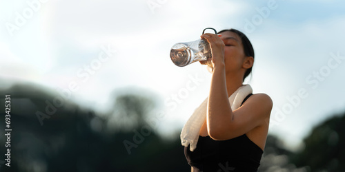 Beautiful young Asian woman stops to drinking water and wipe off her sweat after her evening run at park photo