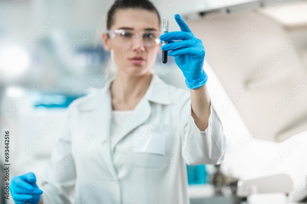 Fototapeta premium Young woman scientist working conducts research in a modern bio laboratory. The doctor holds a test tube with liquid in his hands and examines it