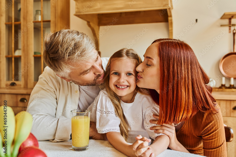 caring parents kissing daughter near fresh fruits and orange juice in kitchen, love and care