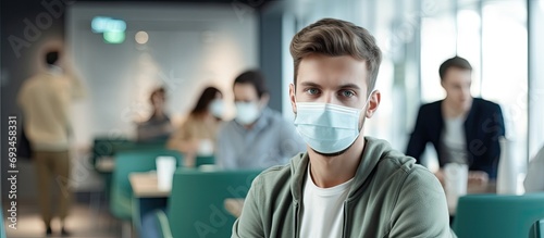 Focus on the young man wearing a face mask, socially distancing in the waiting room of a hospital or office, alongside others of all ages.