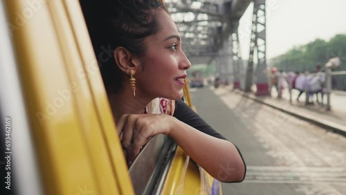 A young happy pretty Indian Asian ethnic Hindu traditional woman or female is sticking her head out of the taxi window to enjoy the view of the iconic Howrah bridge while travelling in a yellow cab