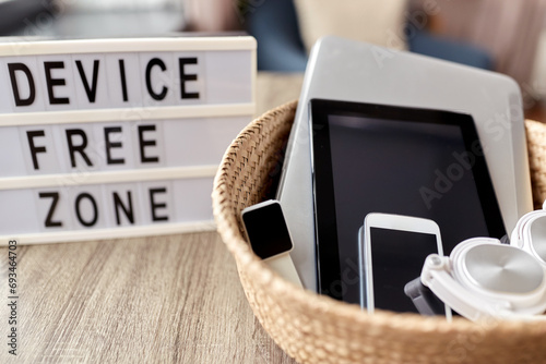 digital detox and technology concept - close up of device free zone words on light box and different gadgets in wicker basket on table at home