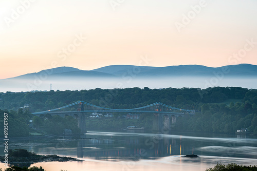 The Menai Bridge, crossing from north Wales to the island of Angelsey, reflected in the water. It is dawn, and a layer of low cloud can be seen shrouding the moutains in the distance © parkerspics